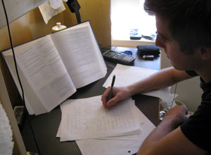 A photograph shows a male student seated at a desk. A book is propped open in front of the student, and he appears to be using the book as a reference as he takes notes on lined paper on the desk in front of him.