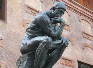 A photograph shows the sculpture entitled The Thinker. The sculpture is of a nude man seated on a rock, his chin resting on his right arm. His right elbow is supported by his left thigh; his left hand is draped over his left knee. He is staring at a point in space and appears to be thinking.